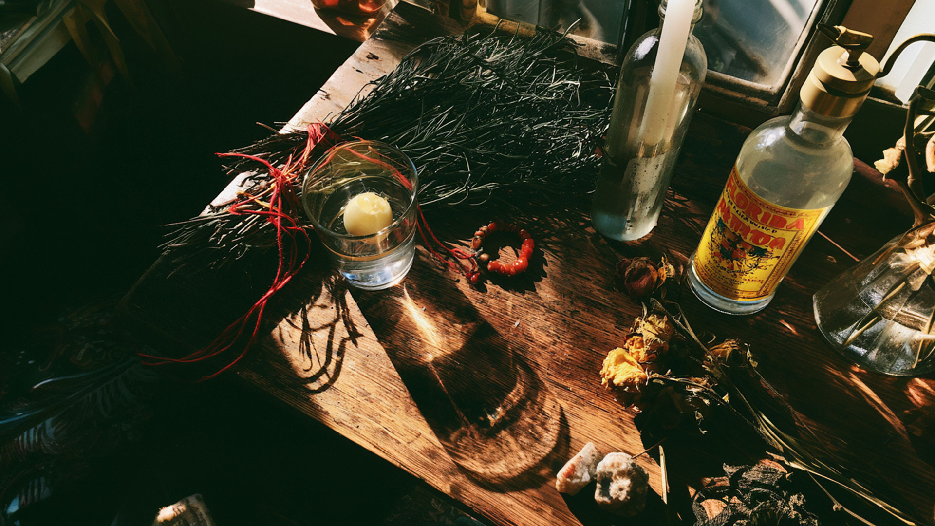 Abuela Rituals: 6 Sacred Cleansings TikTok Stole and Resold concise, descriptive, ends with disclosure): Curandera's altar on a wooden table — glass of water with a cracked egg for limpia, rosemary tied with red string, an azabache bracelet, Florida water, a white candle and dried roses. Illustration, artist's interpretation by Wehpa, created with AI.